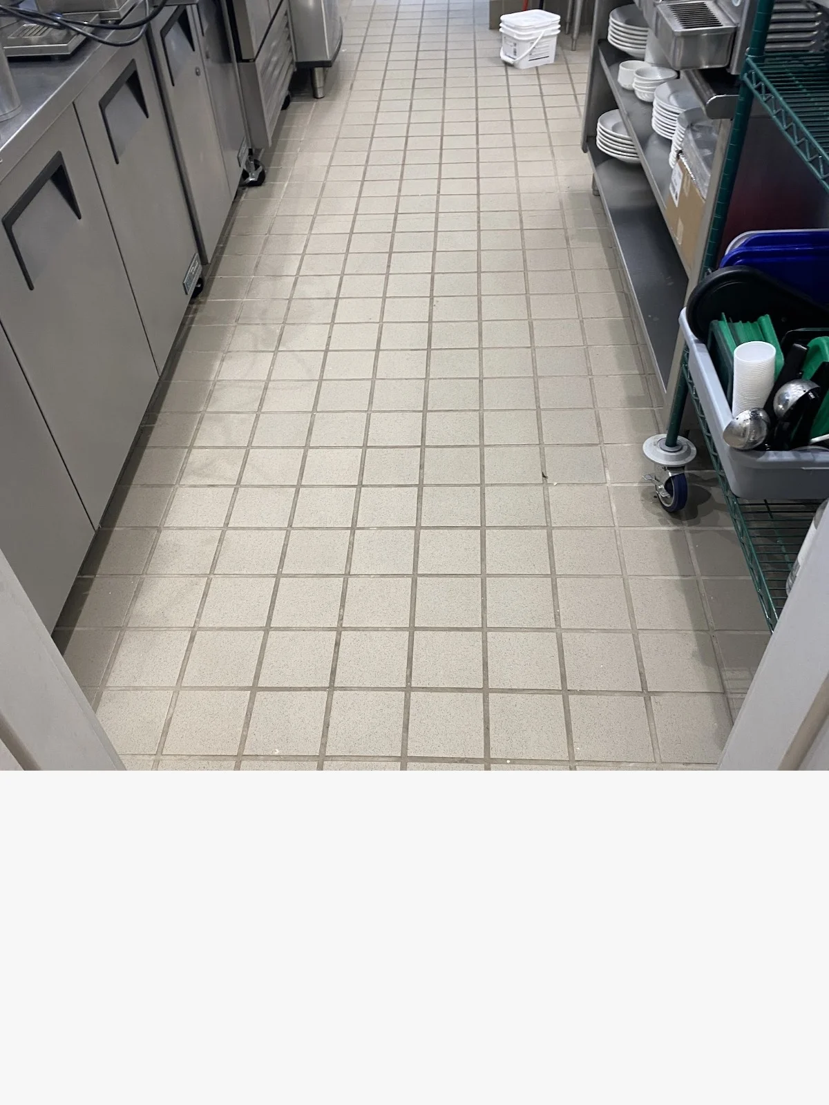 Clean commercial kitchen passageway with tiled floor, stainless steel equipment, and storage containers
